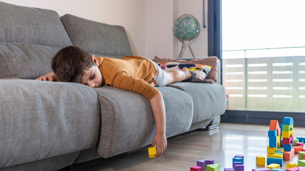 znudzony chłopiec leży na kanapie obok rozrzucone zabawki – symbol nudy i przerwy w zabawie.