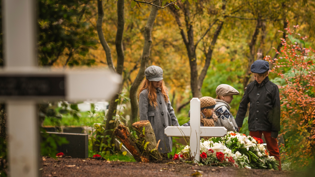 Rodzice z dziećmi odwiedzają grób bliskich w jesienny dzień. Gest czułości i pamięci łączy pokolenia. Rodzinne wspomnienia i rozmowy o bliskich. Dzieci uczą się pamięci przez gesty, obecność i rozmowę. Tak rodzi się tradycja, która trwa przez lata.